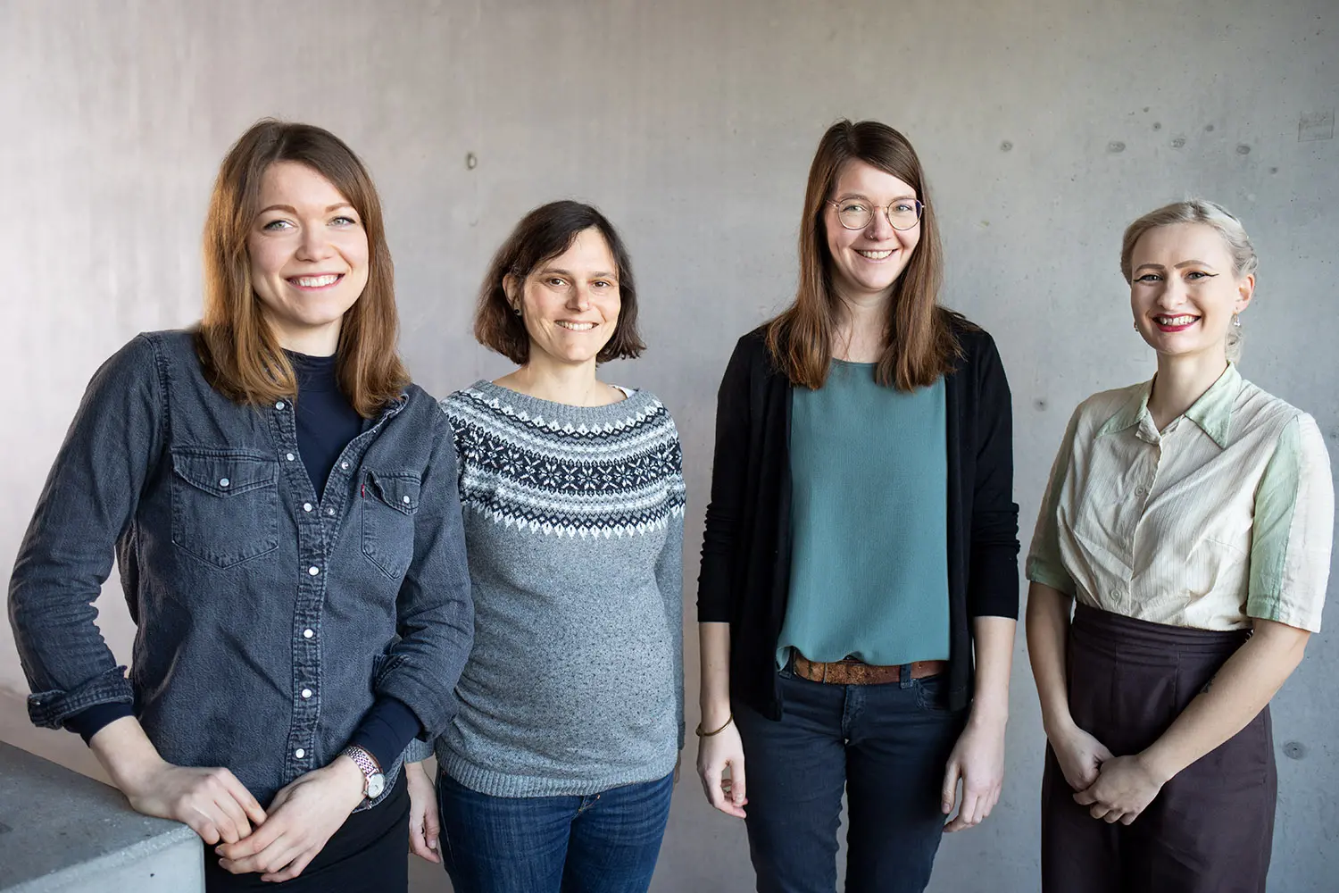 Das Bild zeigt vier Frauen, die lächelnd in die Kamera schauen. Sie stehen nebeneinander und befinden sich vor einer grauen Wand. 