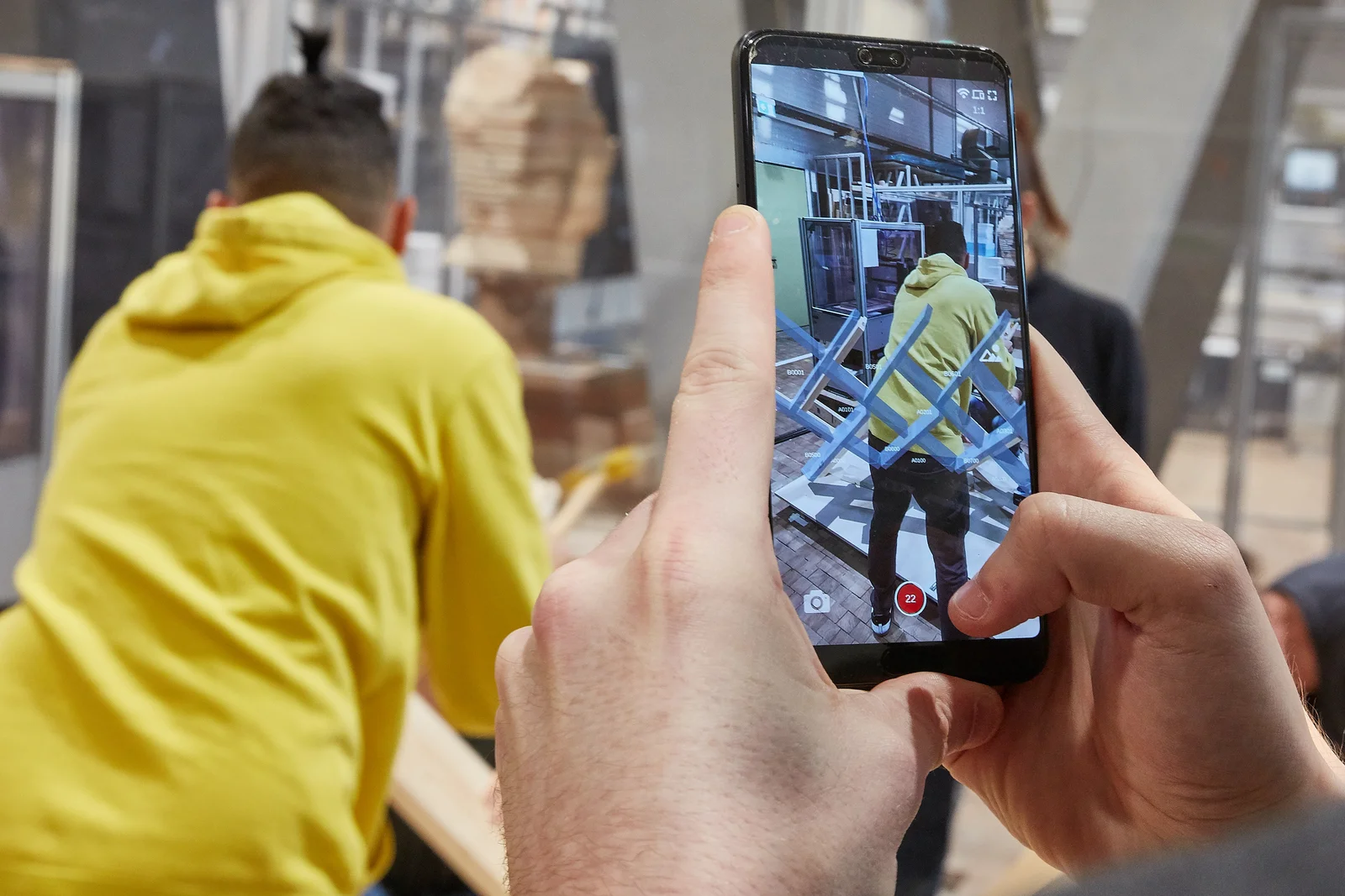Holzbau mit Hilfe von Virtual Reality Blick auf ein Smartphone Display, welches eine Aufbauanleitung für einen Holzaufbau anzeigt. Diese Anleitung ist durchscheinend, so dass auf dem Display ebenfalls eine Person sichtbar ist, welche ein nummeriertes Bauteil an der richtigen Stelle einbaut. Es handelt sich also um eine Art virtuelle Aufbauanleitung für ein Holzständerbauwerk.