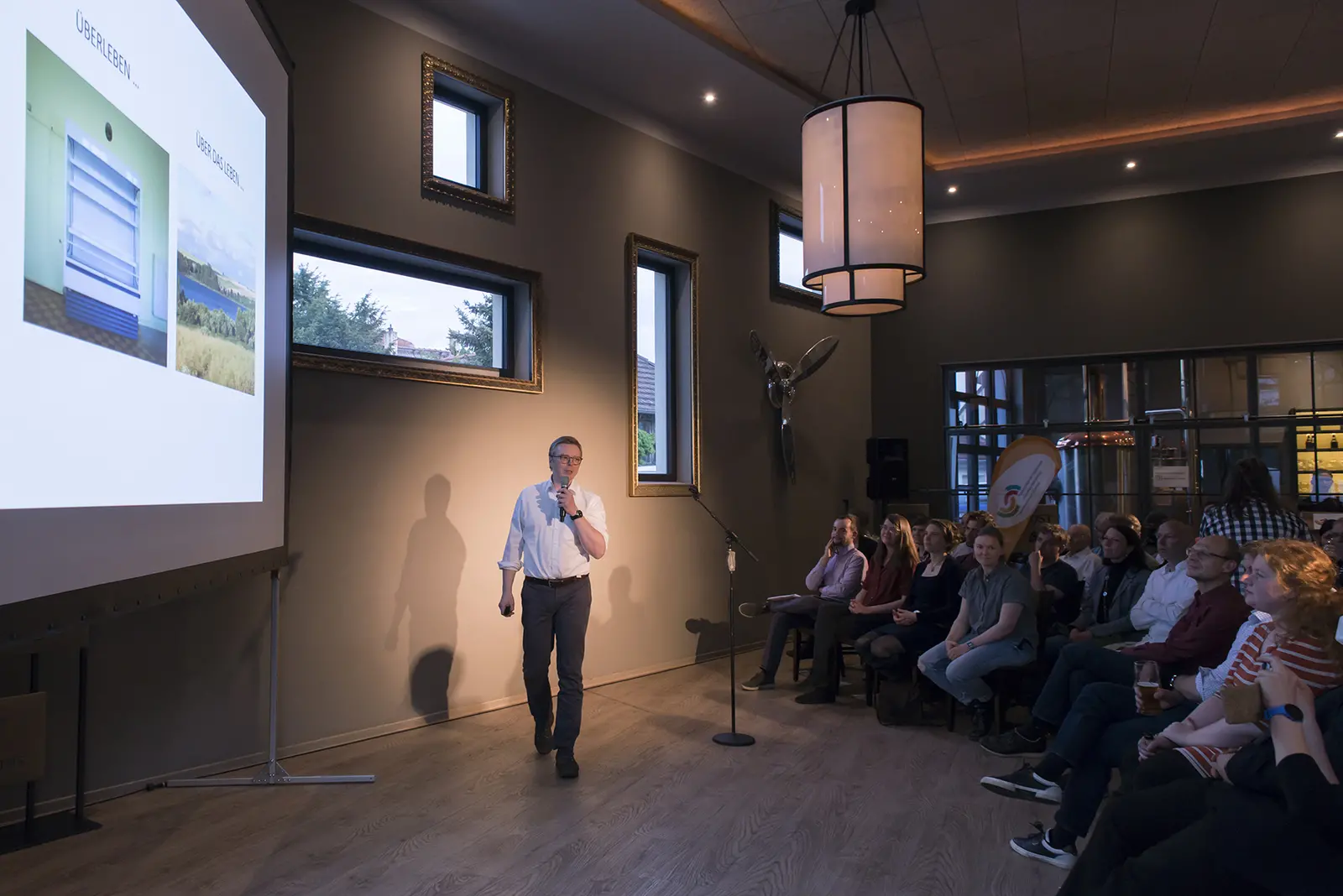 Das Bild zeigt einen Sprecher während des Brandenburger Science Slam in Schwedt. Er steht vor einem Publikum und präsentiert auf einer Leinwand Bilder von zwei unterschiedlichen Landschaften. Die Zuhörer sitzen auf Stühlen und hören aufmerksam zu.