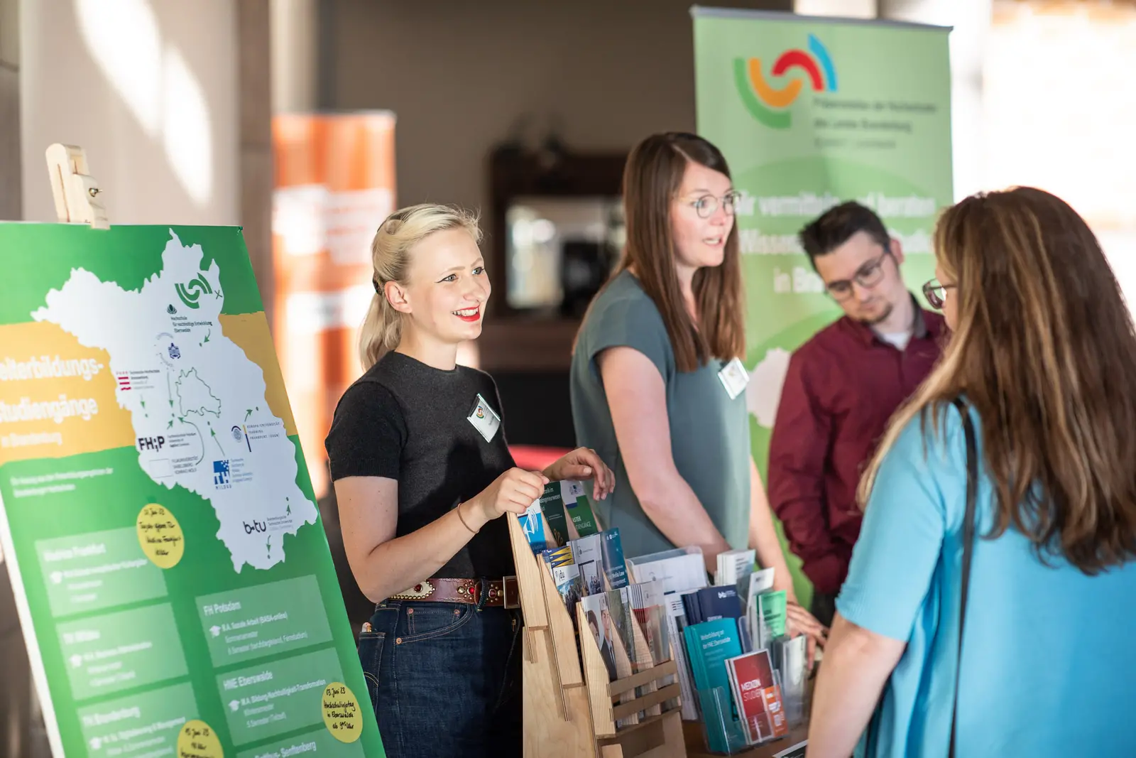 Auf dem Bild ist eine Frau zu sehen, die lächelnd an einem Informationsstand steht. Sie hält Informationsmaterial in der Hand und unterhält sich mit einer Person, die nicht im Bild zu sehen ist. Im Hintergrund befindet sich ein Poster mit einer Landkarte und Informationen über Studienmöglichkeiten in Brandenburg.