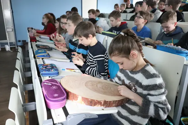 Schulkinder sitzen im Hörsaal und begutachten eine Holzscheibe. Ein Mädchen zählt die Jahrringe.