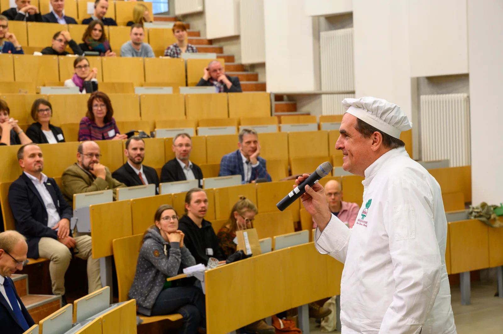 Science meets business Man blickt in einen Hörsaal, welcher gut gefüllt ist. Im Vordergrund steht ein Mann mit einem Mikrofon in der Hand und erzählt etwas. Seine weiße Jacke und die weiße Mütze, die er trägt, zeigen, dass er Bäcker ist.