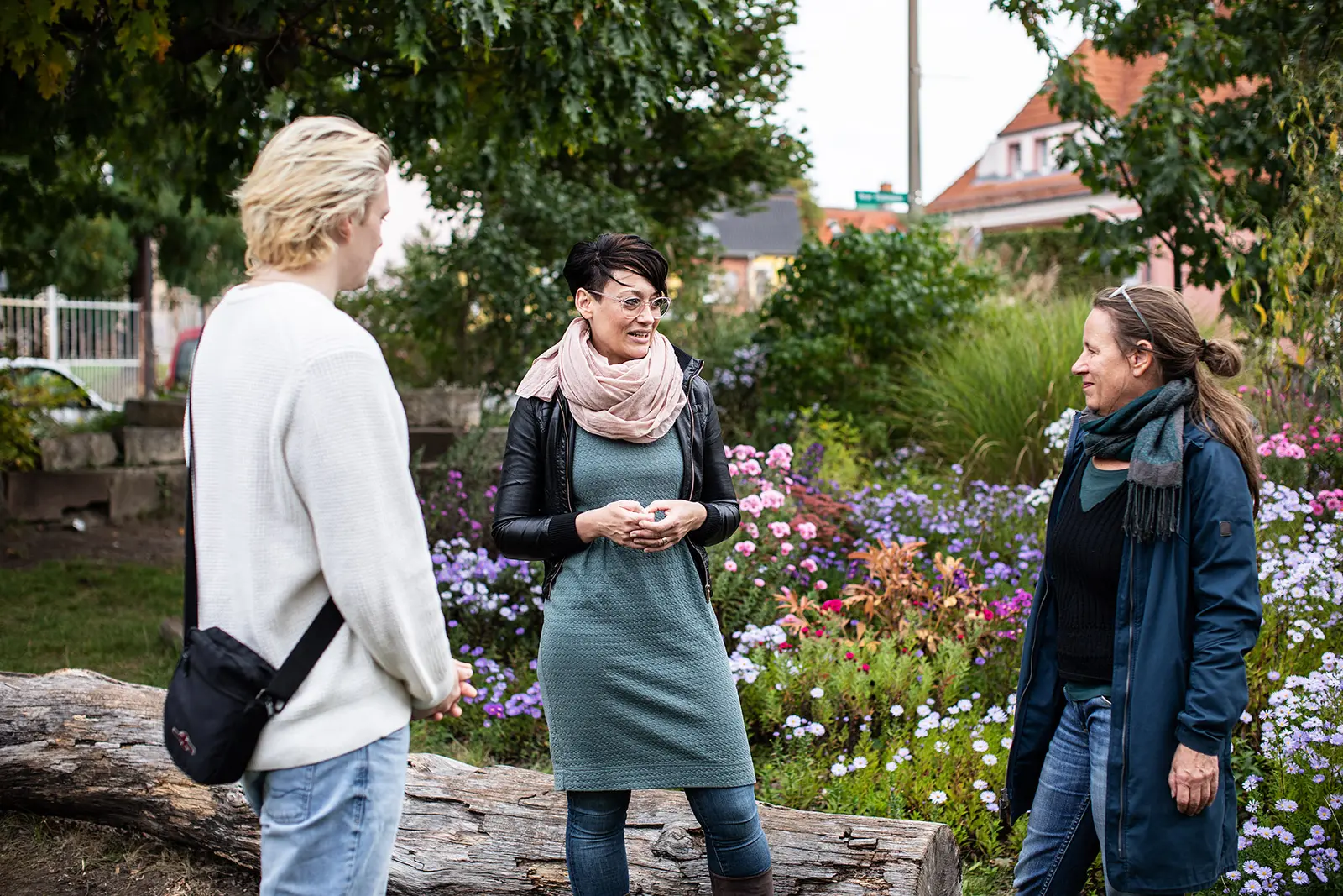 Drei Personen stehen vor einer bunt blühenden mit Stauden bepflanzten und von Bäumen beschatteten Fläche. Im Hintergrund sieht man Häuser. Es handelt sich um den sogenannten Blumenplatz in Eberswalde. Eine Dozentin der Hochschule ist im Gespräch mit einer Partnerin der Zivilgesellschaft und einem Studierenden.