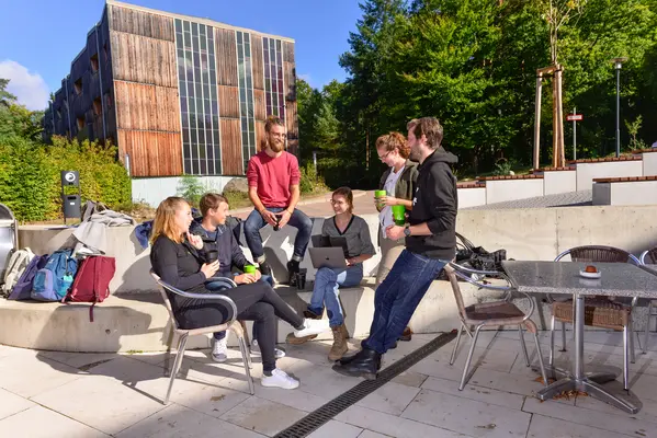 Sechs Studierende sitzen oder stehen um einen Tisch herum und unterhalten sich freudig.